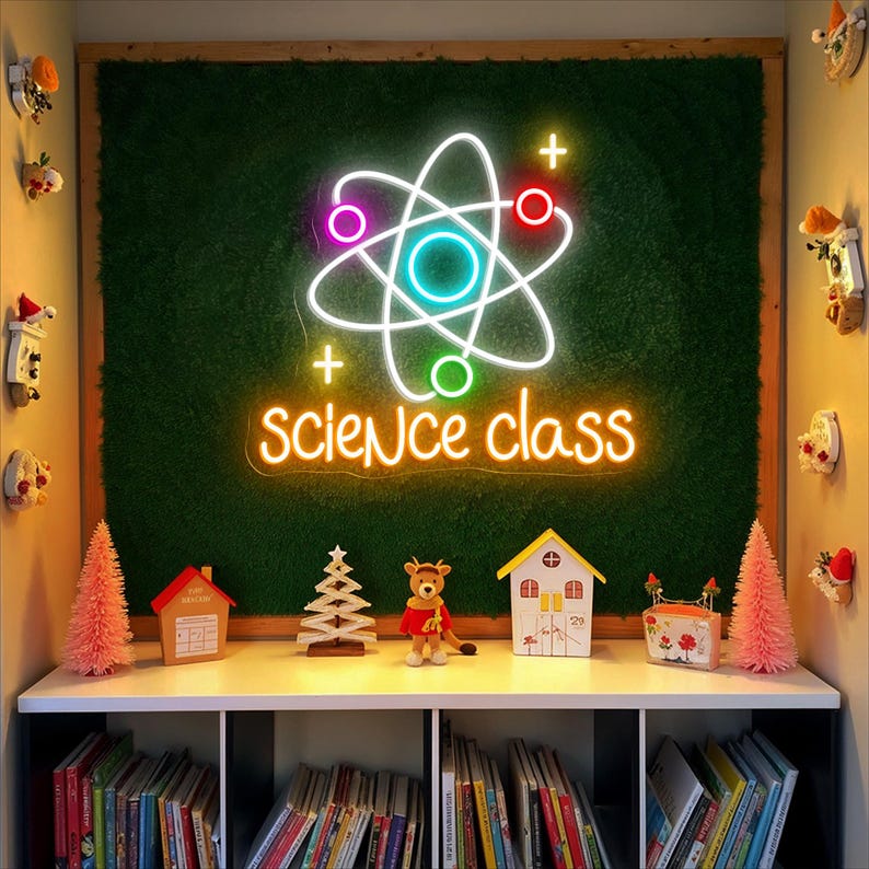 May include: A neon sign with a science theme, featuring an atom symbol and the words "science class" in a bright, glowing font. The sign is mounted on a green textured background. Below, a shelf displays books and decorative items, including miniature houses and a reindeer figurine.