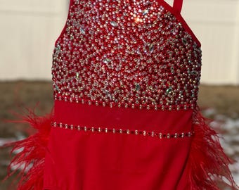 May include: Red dance leotard with a high neckline and a one-shoulder design. The leotard is covered in rhinestones and has a feather trim.