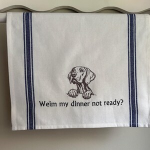 May include: White kitchen towel with blue stripes and a Weimaraner dog illustration. The text "Weim my dinner not ready?" is printed below the dog. The towel is hanging on a white rod.