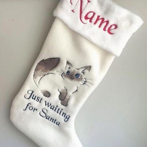 May include: White Christmas stocking with a cute embroidered cat design. The stocking has the text "Just waiting for Santa" and a space for a name.