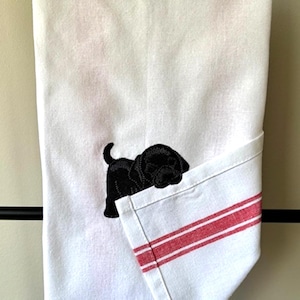 May include: White kitchen towel with a black embroidered Labrador puppy design. The towel has a folded corner with red stripes. The towel is hanging on a black bar.