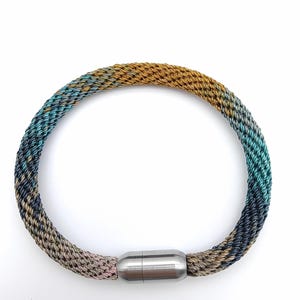 May include: A multi-colored braided necklace with a magnetic clasp. The necklace features a gradient of colors, including gold, teal, blue, and pink. The clasp is a silver-colored metal. The necklace is circular in shape.