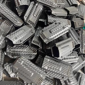 May include: A pile of black and gray carbon fiber holsters. Each holster has a textured, woven pattern and a clip on the back. The holsters have a rectangular shape with a curved top and a series of ventilation slots.