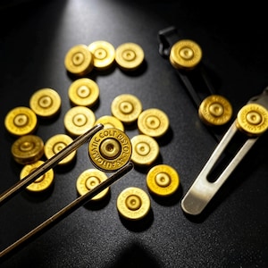 Op de afbeelding: Een verzameling goudkleurige, ronde metalen onderdelen die lijken op kogelhulzen, waarvan er één wordt vastgehouden door een metalen pincet. Elk stuk is voorzien van de tekst 45 AUTOMATIC COLT PISTOL. Rechts bevindt zich een zilverkleurige metalen clip.