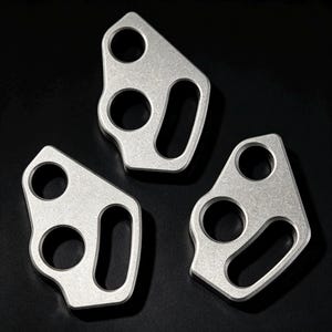 May include: Three identical silver-colored metal components, each featuring a distinct asymmetrical shape with two circular cutouts and one elongated oval slot. The surfaces have a matte, textured finish, and the edges are clearly defined.