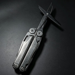 Op de afbeelding: Een zwart multifunctioneel gereedschap met een opvouwbaar ontwerp en uitgeklapte schaar. Het handvat toont de in reliëf aangebrachte tekst LEATHERMAN. Het gereedschap is gemaakt van donker metaal met zichtbare scharnierpunten en grip.
