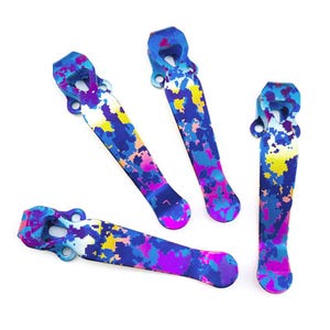 May include: Four metal pocket clips featuring a vibrant digital camouflage pattern in shades of blue, purple, pink, and yellow. Each clip has a curved, elongated shape with a mounting base containing two circular holes for attachment purposes.