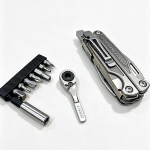 Op de afbeelding: Een zilverkleurige Leatherman Charge multitool met broekclip, een kleine zilverkleurige Fireseed ratelsleutel en een zwarte kunststof bithouder met diverse metalen schroevendraaierbits en een zilverkleurige bitverlengadapter.