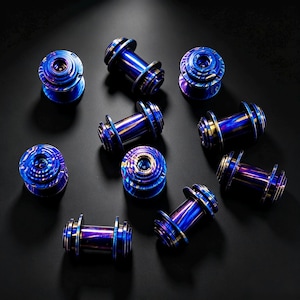 Puede incluir: Un conjunto de nueve componentes metálicos en forma de bobina con un acabado iridiscente en azul y morado vibrante. Cada pieza tiene un eje cilíndrico central con extremos circulares con reborde y una superficie pulida y reflectante.