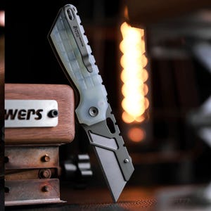 May include: A folding utility knife with a matte grey metal handle and a translucent, textured light-colored grip section. The handle features a metal pocket clip and the text North Mountain Blade. The blade is a straight-edged, flat-ground design.