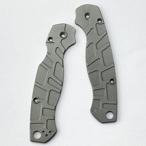 Op de afbeelding: Twee grijze metalen mesheftschalen met een verzonken geometrisch rasterpatroon. Elk onderdeel heeft voorgeboorde ronde gaten voor montageschroeven en een grotere ronde opening aan de basis, afgewerkt met een matte textuur en contouren.