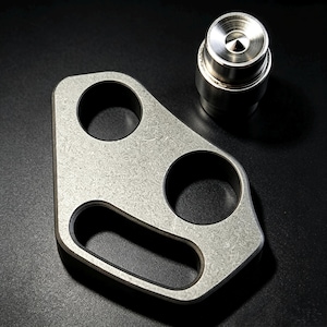 Könnte beinhalten: Ein metallisches, silberfarbenes Werkzeug bestehend aus einer flachen, unregelmäßig geformten Platte mit zwei kreisförmigen Löchern und einem länglichen ovalen Schlitz, begleitet von einem separaten, zylindrischen Metallteil mit einer spitzen, konischen Vertiefung.