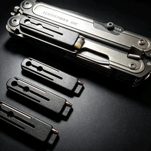 Pode incluir: Uma ferramenta multifunções Leatherman ARC em posição fechada com acabamento prateado e preto, apresentando o texto LEATHERMAN ARC e USA. Três clipes de bolso de metal preto estão dispostos em fila abaixo da ferramenta numa superfície escura.