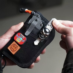 Puede incluir: Funda EDC (Every Day Carry) de nailon negro con un clip para cuchillo en forma de calavera y dos navajas. La funda tiene dos parches: uno rojo con una calavera y el texto "MacArmy", y otro marrón con un diseño de "X".