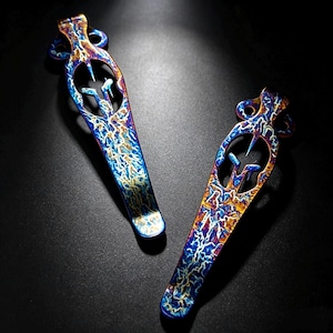 May include: Two elongated metal pocket clips featuring a vibrant, heat-anodised finish with swirling patterns of blue, gold, and purple. Each clip has a decorative cutout design at the top end, resembling a stylised helmet or crest shape.