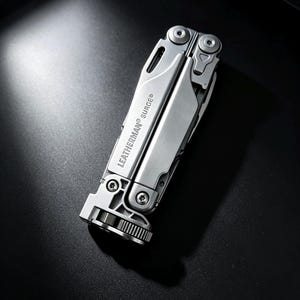 Könnte beinhalten: Ein silbernes Leatherman Surge Multifunktionswerkzeug in geschlossener, zusammengeklappter Position. Der Edelstahlkörper weist sichtbare Schrauben, einen texturierten Verriegelungsmechanismus an der Basis und den Markennamen Leatherman Surge auf.