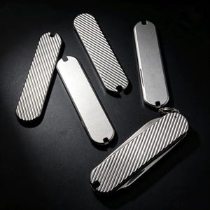 Pode incluir: Um conjunto de cinco talas de metal prateado para canivete, incluindo três com textura de estrias diagonais e duas com acabamento liso. Uma tala está fixada a um canivete dobrado que possui uma pequena argola de metal na extremidade.