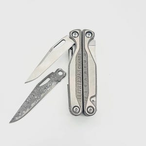 May include: A silver Leatherman Charge Titanium multi-tool with one blade deployed, marked S30V. Beside it lies a separate, detached knife blade featuring a distinct, intricate Damascus steel pattern with a circular hole near the base.