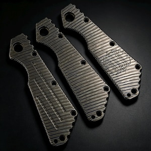 May include: Three metallic knife handle scales featuring a dark, weathered finish with a distinct diagonal grooved texture. Each piece includes circular cutouts for assembly hardware, with the rightmost scale displaying a faint circular emblem.