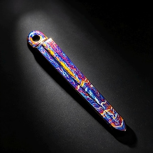 Puede incluir: Una herramienta de metal larga, delgada y plana que presenta un orificio circular en el extremo superior. La superficie muestra un acabado anodizado vibrante y multicolor con patrones de remolinos en tonos azul, naranja, amarillo y violeta.