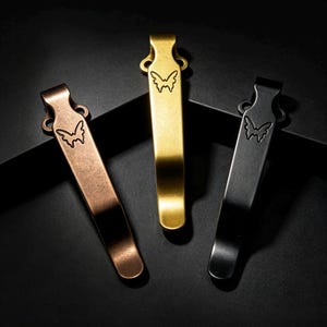 May include: Three metal pocket clips in copper, gold, and black finishes. Each clip features a curved design with two circular mounting holes at the top and a small butterfly icon engraved near the attachment point on the front surface.