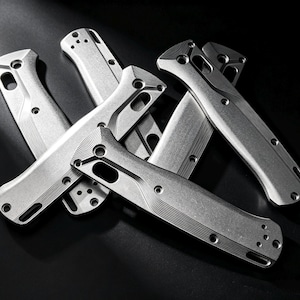 Op de afbeelding: Een verzameling zilverkleurige metalen handgrepen voor messen met een textuur van groeven. Elk onderdeel heeft voorgeboorde gaten voor montage, een voorgevormde structuur en een matte afwerking, gerangschikt in een hoop op een donkere ondergrond.