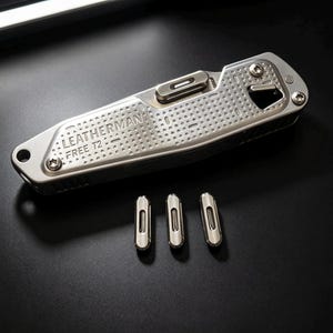 Könnte beinhalten: Ein silbernes Leatherman Free T2 Multifunktionswerkzeug mit strukturiertem Griff und Verriegelungsmechanismus, begleitet von drei kleinen, passenden zylindrischen Metallkomponenten, die in einer Reihe unter dem Werkzeug liegen.