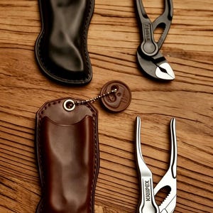 Könnte beinhalten: Zwei Lederscheiden, eine schwarz und eine braun, jeweils mit einer Metallöse und einem runden, mit einem Totenkopf geprägten Anhänger an einer Kugelkette. Neben jeder Scheide liegt eine Knipex-Zangenschlüssel, eine in mattschwarz und eine in Silber.