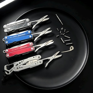 Puede incluir: Cuatro multiherramientas Leatherman Squirt PS4 en negro, azul, rojo y plata, cada una con tijeras desplegadas. Las herramientas están dispuestas junto a varios tornillos metálicos pequeños, pasadores y un componente metálico suelto sobre una superficie oscura.