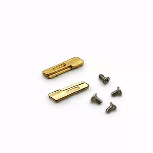 Könnte beinhalten: Zwei rechteckige, goldfarbene Metallplatten mit einem eingelassenen Schlitz auf einer Seite, begleitet von vier kleinen, silberfarbenen Metallschrauben mit Gewindeschaft und Flachkopf, angeordnet auf einer weißen Oberfläche.