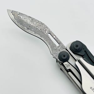 Op de afbeelding: Een inklapbaar zakmes met een gebogen lemmet voorzien van een opvallend zilver- en grijskleurig damaststaalpatroon. Het handvat is opgebouwd uit metalen onderdelen met zwarte ronde scharnierschroeven en een zilveren broekclip aan de zijkant.