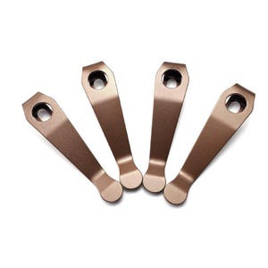 Puede incluir: Cuatro clips de bolsillo metálicos de color bronce dispuestos en forma de abanico. Cada clip presenta un diseño cónico con una punta redondeada y un orificio circular en el extremo superior más ancho, hecho de metal con acabado mate.