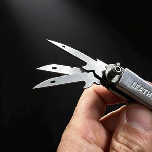 May include: A silver-coloured multi-tool featuring three thin, pointed metal blades with small rectangular cutouts. The tool is constructed from brushed stainless steel and displays the partial text LEATH on the handle, secured by a star-shaped screw.