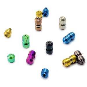 Peut inclure: Une collection de petits composants de quincaillerie métalliques cylindriques dans diverses finitions métalliques, notamment or, argent, bronze, noir, bleu, vert et violet. Chaque pièce présente une extrémité filetée et une tête à empreinte étoilée.