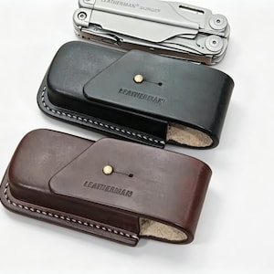 May include: A silver Leatherman Surge multi-tool positioned above two leather sheaths, one black and one dark brown. Each sheath features a brass stud closure, white contrast stitching, and the Leatherman brand name embossed on the front flap.