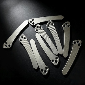 May include: A collection of nine metallic pocket knife handle scales, each featuring a curved, elongated shape with three circular mounting holes at one end. Some scales have a textured, grooved surface pattern, while others have a smooth, matte finish.
