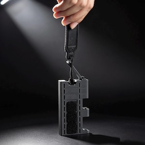 May include: A black rectangular tactical sheath with a textured surface and a central hook-and-loop fastener strip. The sheath is suspended by a black cord attached to a matching black pull tab, featuring a rigid construction with side cutouts.