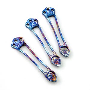 May include: Three elongated metal components with a vibrant, iridescent finish featuring swirling patterns of blue, purple, and gold. Each piece has a triangular head with three circular holes and a pointed, shield-shaped base with a textured design.