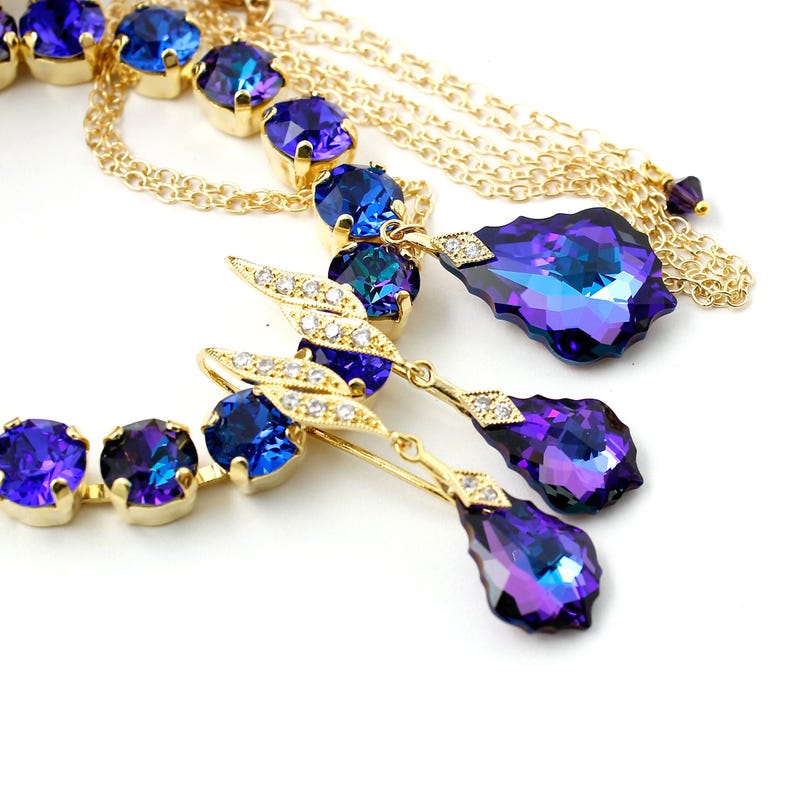 Pode incluir: Um conjunto de colar e brincos de ouro com pedras de cristal iridescentes azuis e roxas. O colar tem uma corrente de v&aacute;rias camadas e os brincos s&atilde;o do tipo pendente.