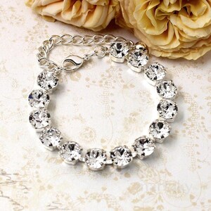 May include: A silver bracelet featuring clear, round-cut crystals set in a linked chain. The bracelet has a lobster clasp closure and is displayed on a patterned surface with yellow flowers in the background.