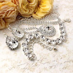 May include: A collection of silver-toned jewelry featuring sparkling clear crystals. The set includes a necklace, bracelet, and earrings. Yellow roses are in the background, adding a touch of color and elegance to the display.