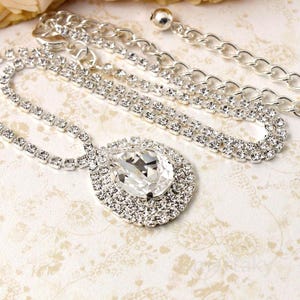 May include: A silver-toned necklace with a large, pear-shaped, clear crystal pendant. The pendant is surrounded by smaller, clear crystals. The necklace features multiple strands of small, clear crystals and a chain.
