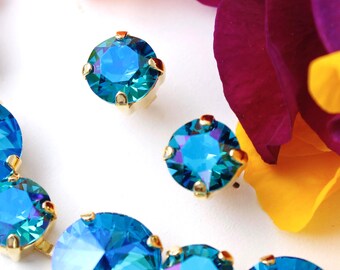 May include: Close-up of several blue and gold-toned crystal earrings. The crystals have a faceted design, reflecting light with shades of blue and green. The earrings are set in gold-coloured metal and are surrounded by colourful flower petals.