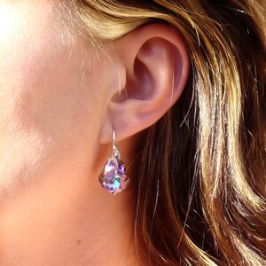 May include: A silver earring with a purple iridescent crystal dangle. The earring is worn in a woman's left ear.