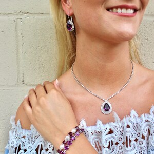 May include: A jewelry set featuring a necklace, earrings, and bracelet. The necklace and earrings have a teardrop-shaped purple gemstone surrounded by clear stones. The bracelet has purple gemstones and white pearls. The jewelry is set against a light blue lace top.