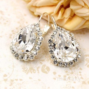 May include: A pair of silver-toned drop earrings featuring large, clear, teardrop-shaped crystals. Each earring is surrounded by a halo of smaller, sparkling crystals. The earrings are set against a soft, floral-patterned background.