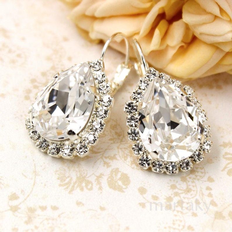 May include: A pair of silver-toned drop earrings featuring large, clear, teardrop-shaped crystals. Each earring is surrounded by a halo of smaller, sparkling crystals. The earrings are set against a soft, floral-patterned background.