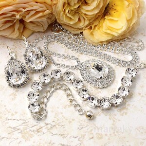 May include: A collection of sparkling silver jewelry, including a necklace with a large teardrop pendant, matching earrings, and a bracelet. The jewelry is adorned with clear, faceted crystals. Yellow roses are in the background.