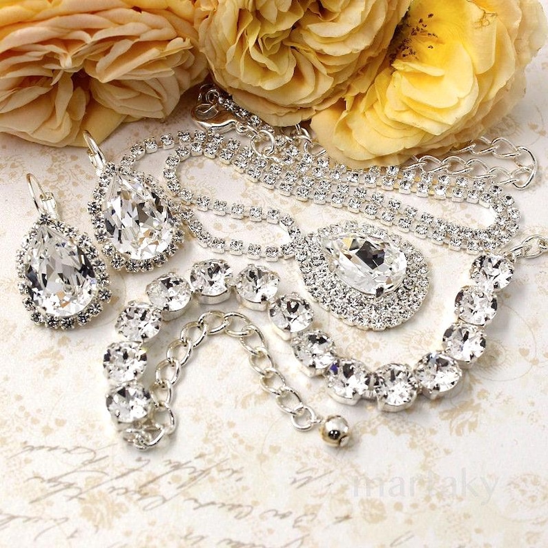 May include: A collection of sparkling silver jewelry, including a necklace with a large teardrop pendant, matching earrings, and a bracelet. The jewelry is adorned with clear, faceted crystals. Yellow roses are in the background.