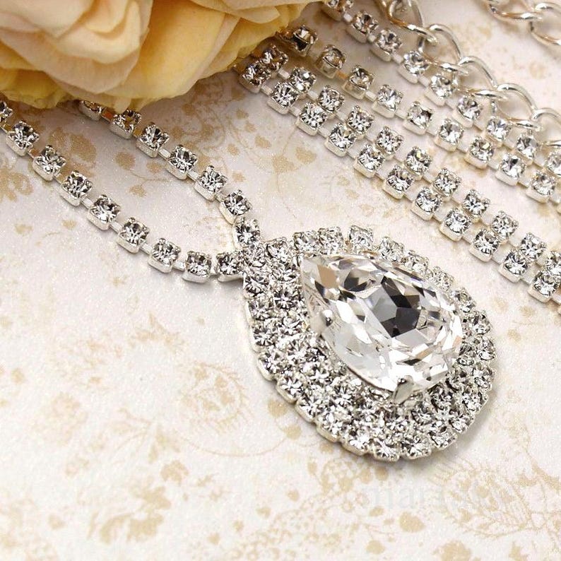 May include: A silver-toned necklace with a large, teardrop-shaped pendant. The pendant and necklace are encrusted with numerous clear, sparkling rhinestones. The background features a floral pattern and a soft, yellow flower.